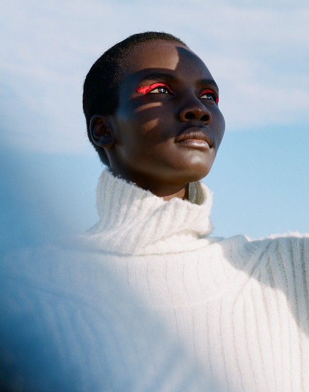 A striking close-up of an African-descent woman with dramatic red eye makeup wearing a white turtleneck against a light blue sky.