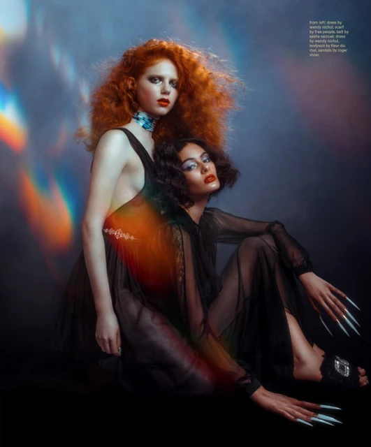 Two women pose in sheer black dresses with long nails. One has red curly hair, the other dark curly hair, against a dark background.