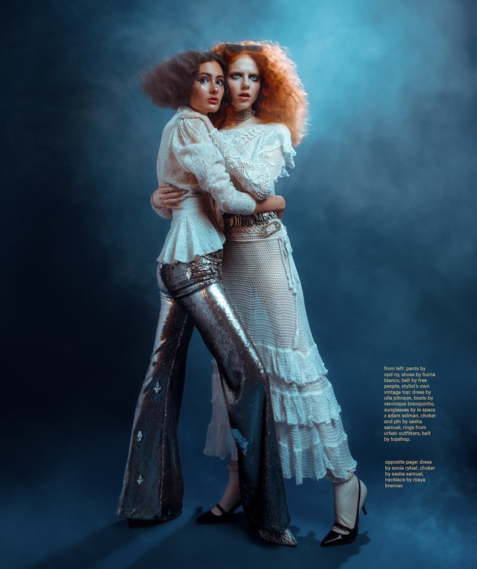 Two fashion models embrace in a studio setting, wearing textured white tops and sequined and ruffled skirts with moody lighting.