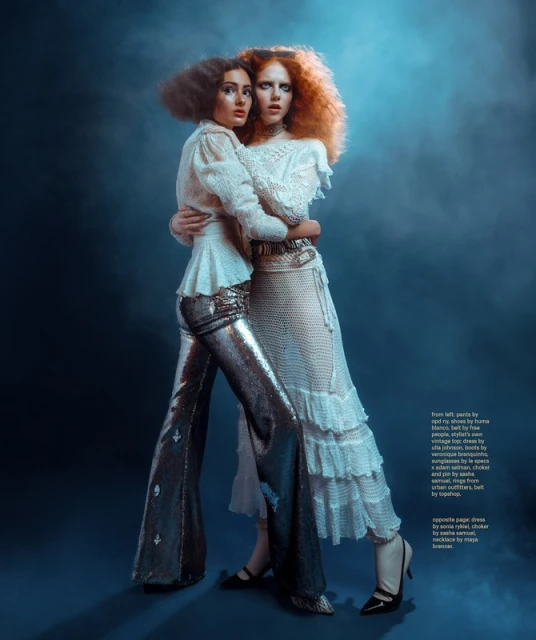 Two fashion models embrace in a studio setting, wearing textured white tops and sequined and ruffled skirts with moody lighting.