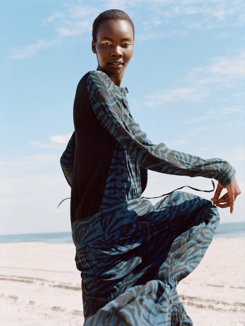 Elegant Black woman dances in a flowing blue zebra-print dress on a beach with a blue sky backdrop, showcasing fashion and freedom.
