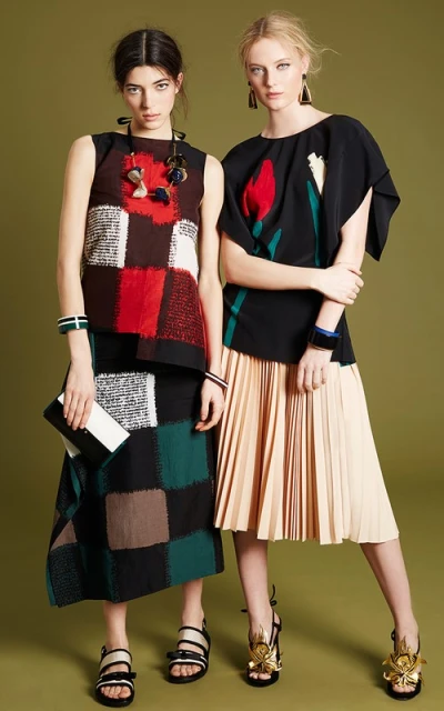 Two women model patterned tops and pleated skirts in a fashion studio, showcasing stylish apparel and accessories against a green backdrop.