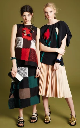Two women model patterned tops and pleated skirts in a fashion studio, showcasing stylish apparel and accessories against a green backdrop.