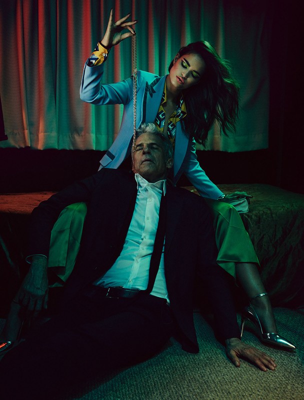 Fashion shot of adult woman holding chain over senior man seated on floor, set in a room with green walls and an olive-green bed.