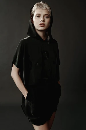 Portrait of a model with short blonde hair wearing a black hooded top against a dark background