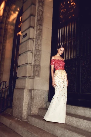 Model standing on stone steps outside an ornate building wearing an off the shoulder beaded gown with red and white detailing.