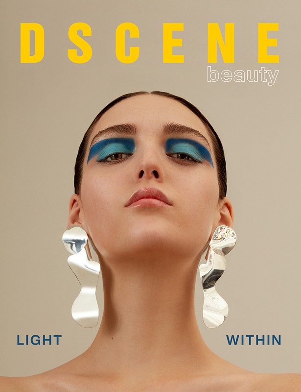 Close-up beauty shot of a young woman with bright blue eye makeup, large silver earrings, and the word 'DSCENE' above her head.