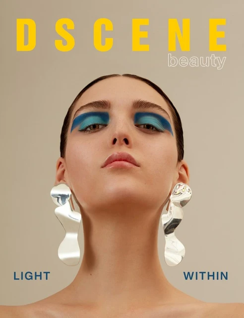 Close-up beauty shot of a young woman with bright blue eye makeup, large silver earrings, and the word 'DSCENE' above her head.