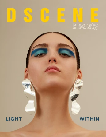 Close-up beauty shot of a young woman with bright blue eye makeup, large silver earrings, and the word 'DSCENE' above her head.