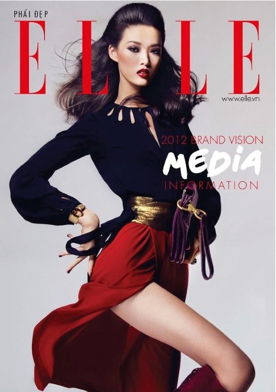 Fashion editorial shot of a female model with red lipstick wearing a black top, gold belt, and red skirt against a light gray background.