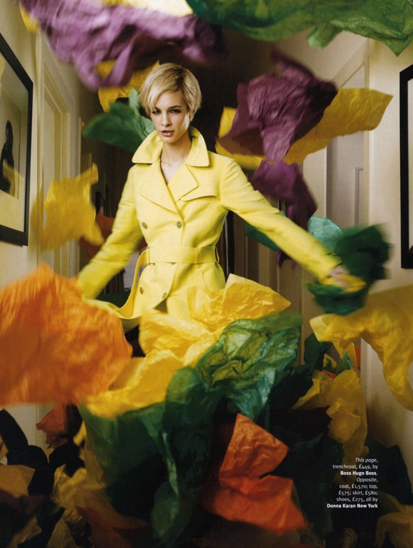 Fashion editorial shot of a woman in a yellow trench coat surrounded by colorful tissue paper indoors, in a magazine-style setting.
