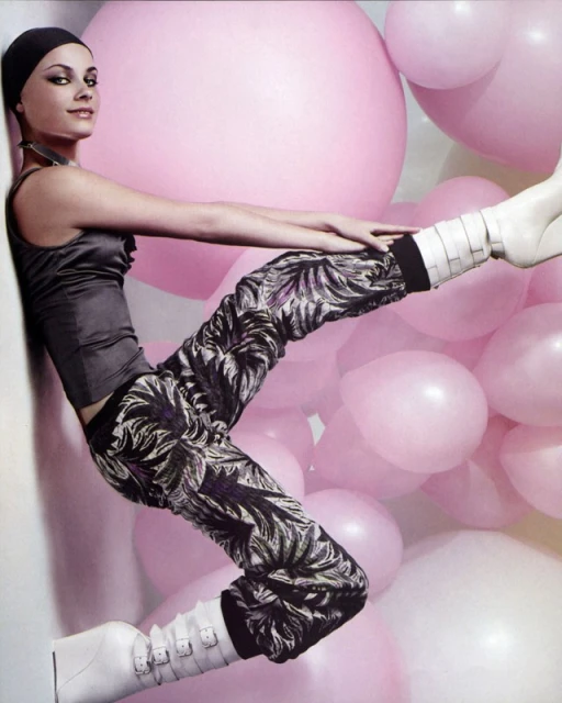 A fashion model strikes a pose with a backdrop of pink balloons while wearing an ash-grey satin top, patterned pants, and white boots.