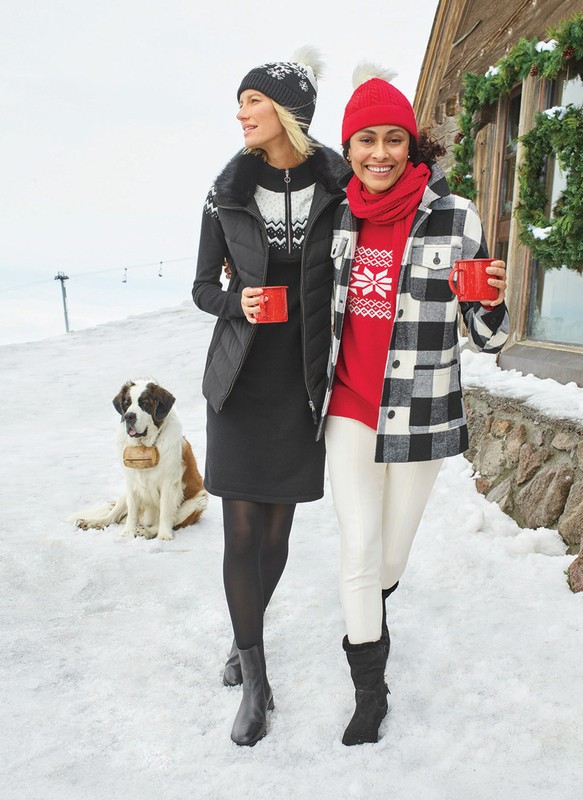 Two adult females in winter wear walk outside on snow-covered ground with a large dog, wearing hats, and jackets while holding mugs.