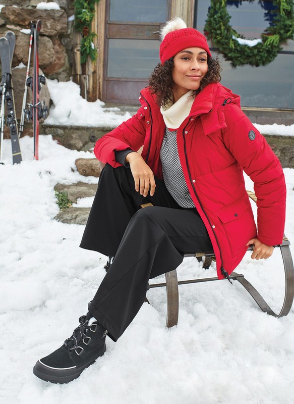 A woman in a red coat and black pants sits on a sled in the snow outside a stone house with skis and a wreath visible.