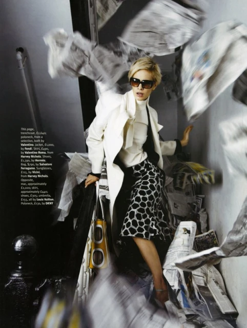 A blonde woman in a white jacket and patterned skirt stands surrounded by flying newspaper pages in a fashion editorial shot.