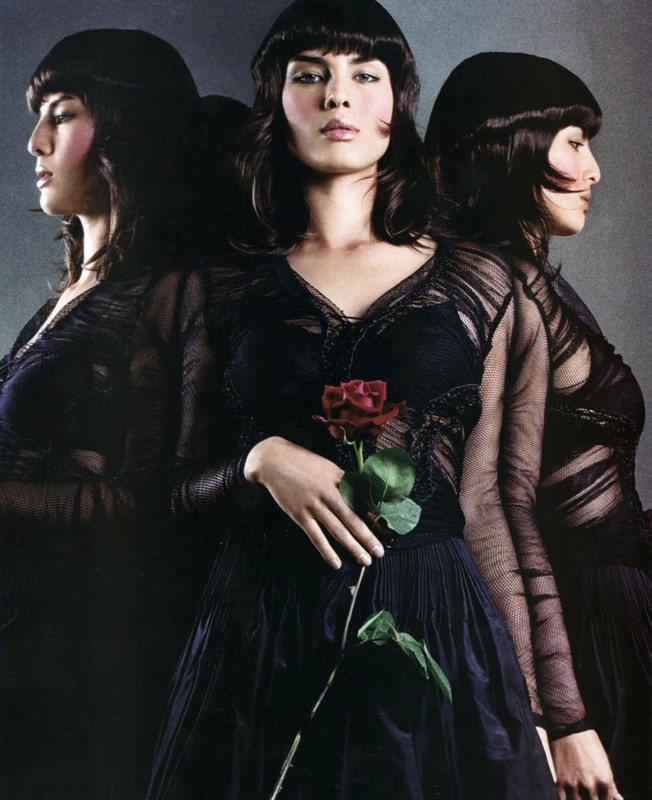 Fashionable woman in black dress holding a red rose, reflected in mirrors, creating a triple portrait against a gray background.