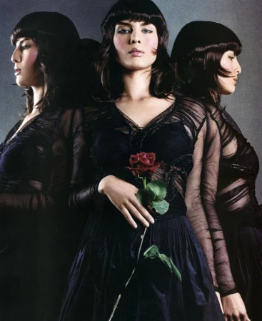 Fashionable woman in black dress holding a red rose, reflected in mirrors, creating a triple portrait against a gray background.