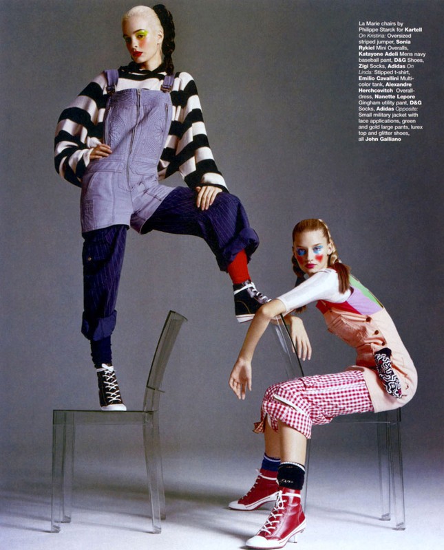 Two fashion models pose with a playful, retro vibe; one stands on a clear chair wearing overalls and a striped top; the other sits.