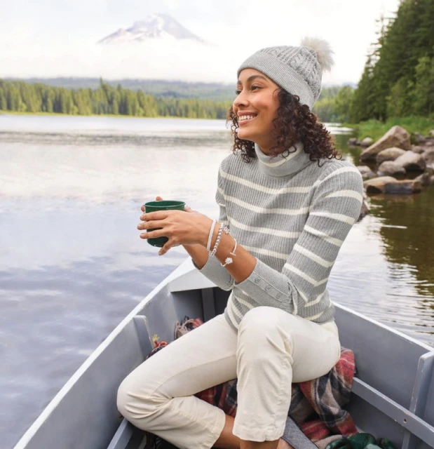 Woman in a grey sweater and beanie sitting in a rowboat on a lake with mountains in the background, holding a mug and smiling.