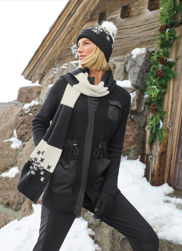 Adult woman in winter clothing featuring a jacket, hat, and scarf posing outdoors against a stone wall and wood cabin, in daylight.