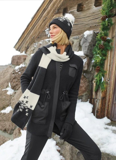 Adult woman in winter clothing featuring a jacket, hat, and scarf posing outdoors against a stone wall and wood cabin, in daylight.