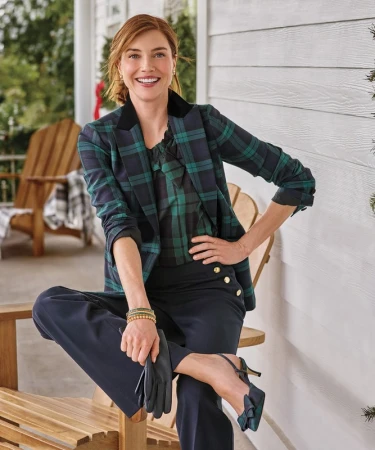 Adult woman in plaid blazer and black pants poses on porch with wood chairs, showcasing fashion in an outdoor setting with a smile.