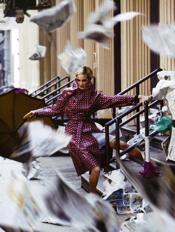 Fashion editorial shot of a woman in a polka dot raincoat on outdoor steps surrounded by flying newspapers, holding an umbrella.