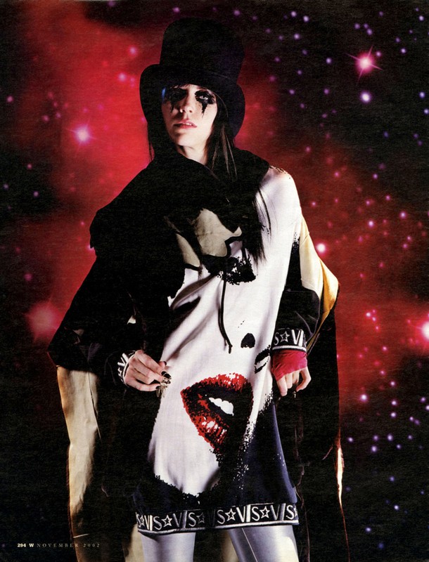 Person with dark eye makeup wearing a top hat and sweater with a large lip print stands against a galactic starburst backdrop.
