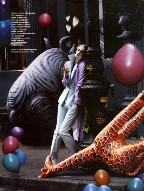 Fashion editorial featuring a woman in a lavender suit, leaning against a Parisian column amidst inflatable animal decorations and balloons.