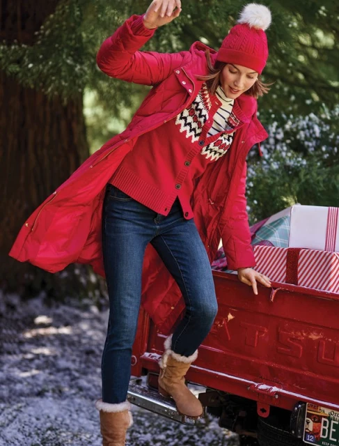A woman in a red coat and hat is stepping onto the bumper of a red pickup truck with presents, surrounded by snowy trees.
