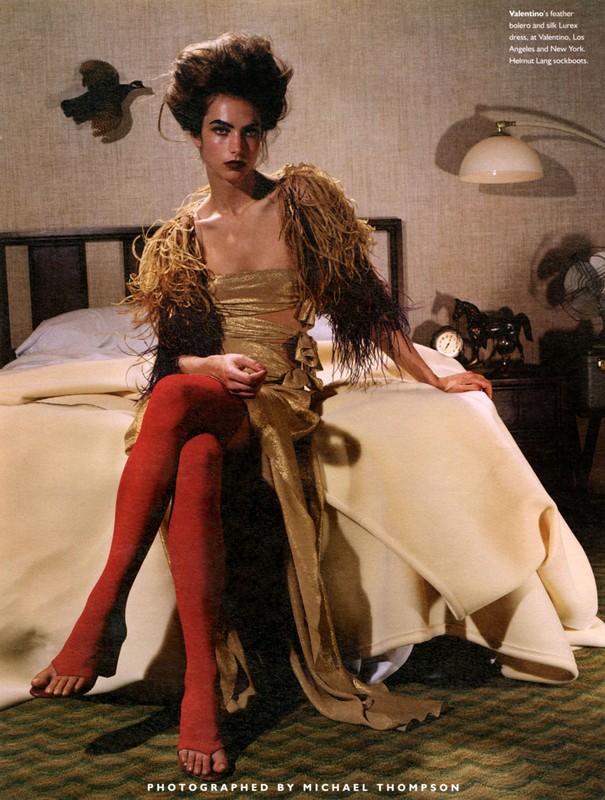 Fashion editorial shot featuring a woman in gold dress and red stockings sitting on a bed in a bedroom setting.