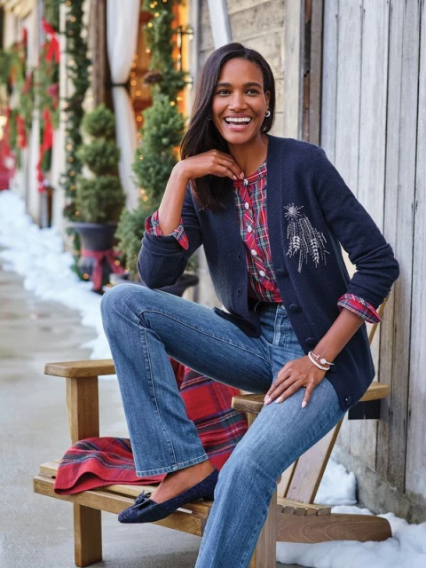 Adult woman in a navy cardigan and jeans sitting on a wooden chair outdoors near greenery and a wood building with holiday decor.