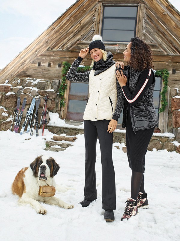 Two women pose with a Saint Bernard dog in a snowy, mountainous landscape, with a wooden cabin and skis in the background. 