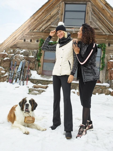 Two women pose with a Saint Bernard dog in a snowy, mountainous landscape, with a wooden cabin and skis in the background. 