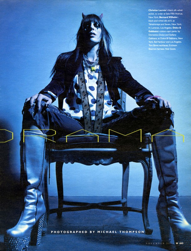 Fashion editorial shot of a slender adult woman with devil horns, sitting in a chair, and wearing a jacket and platform boots.