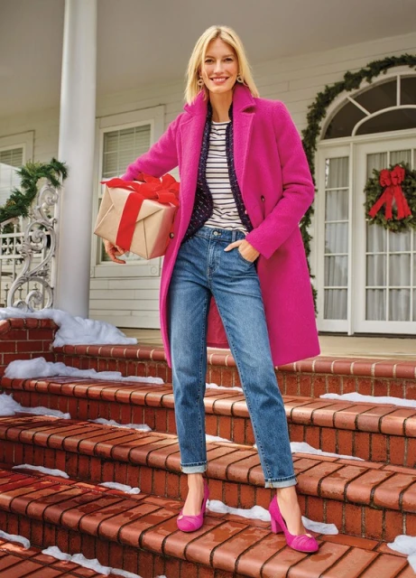 Smiling blonde woman in a pink coat stands on red brick steps with a wrapped gift in her arms, with a white house in the background.