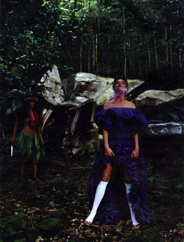 Two women in an outdoor wooded area with airplane wreckage, one wearing a purple dress, the other in native dress with a knife.