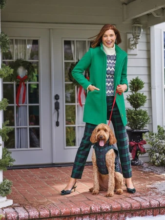 Woman in green coat with Goldendoodle dog wearing vest, standing on porch with Christmas decorations by front door.