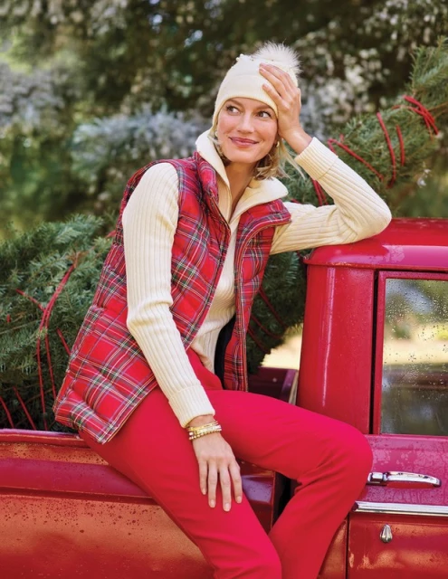 Smiling woman sits on a vintage red pickup truck wearing a red plaid vest, cream sweater, red pants and winter hat, holding a Christmas tree.