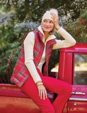 Smiling woman sits on a vintage red pickup truck wearing a red plaid vest, cream sweater, red pants and winter hat, holding a Christmas tree.