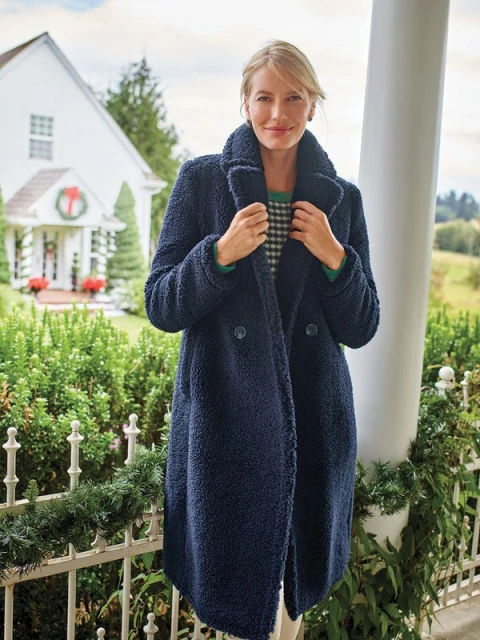 Woman in navy teddy coat standing on porch of white house with Christmas wreath. Outdoor fashion portrait on overcast day.