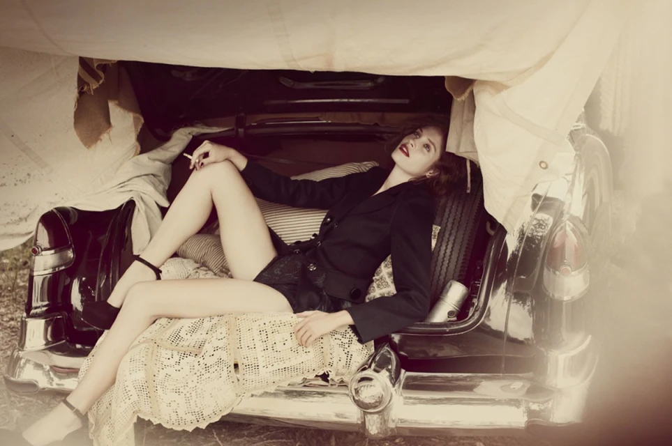 Woman lounges in a vintage car trunk with a cigarette, adorned with pillows, lace, and a canvas cover, blending retro glamour with automotive charm.