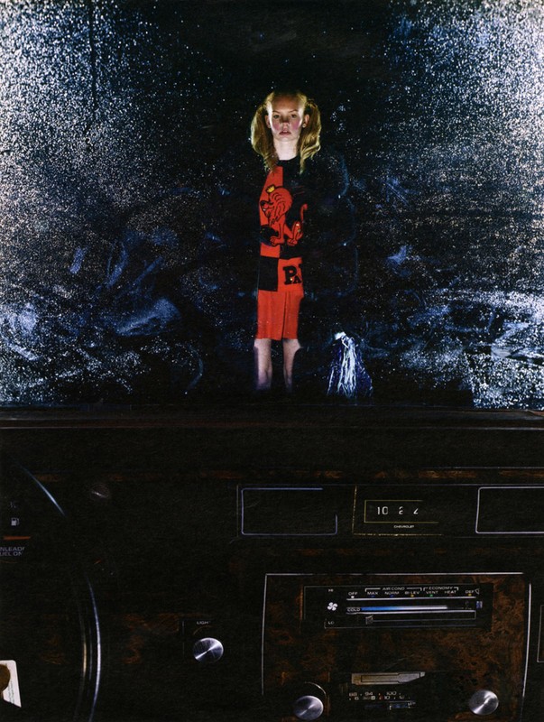 A young child stands in the rain behind a car windshield, viewed from inside the vehicle with a dark, moody atmosphere.