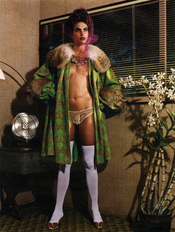 Woman in patterned fur-trimmed coat, lace lingerie, and white stockings poses in a room with blinds, flowers, and a vintage fan.