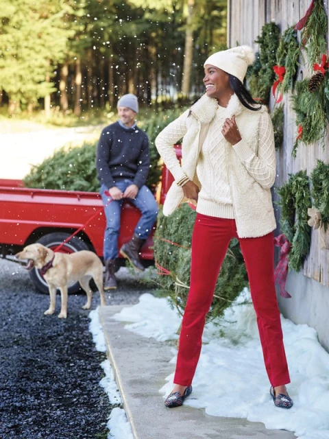 A woman in a winter outfit standing near a red truck with a Christmas tree and a man sitting in the truck bed.