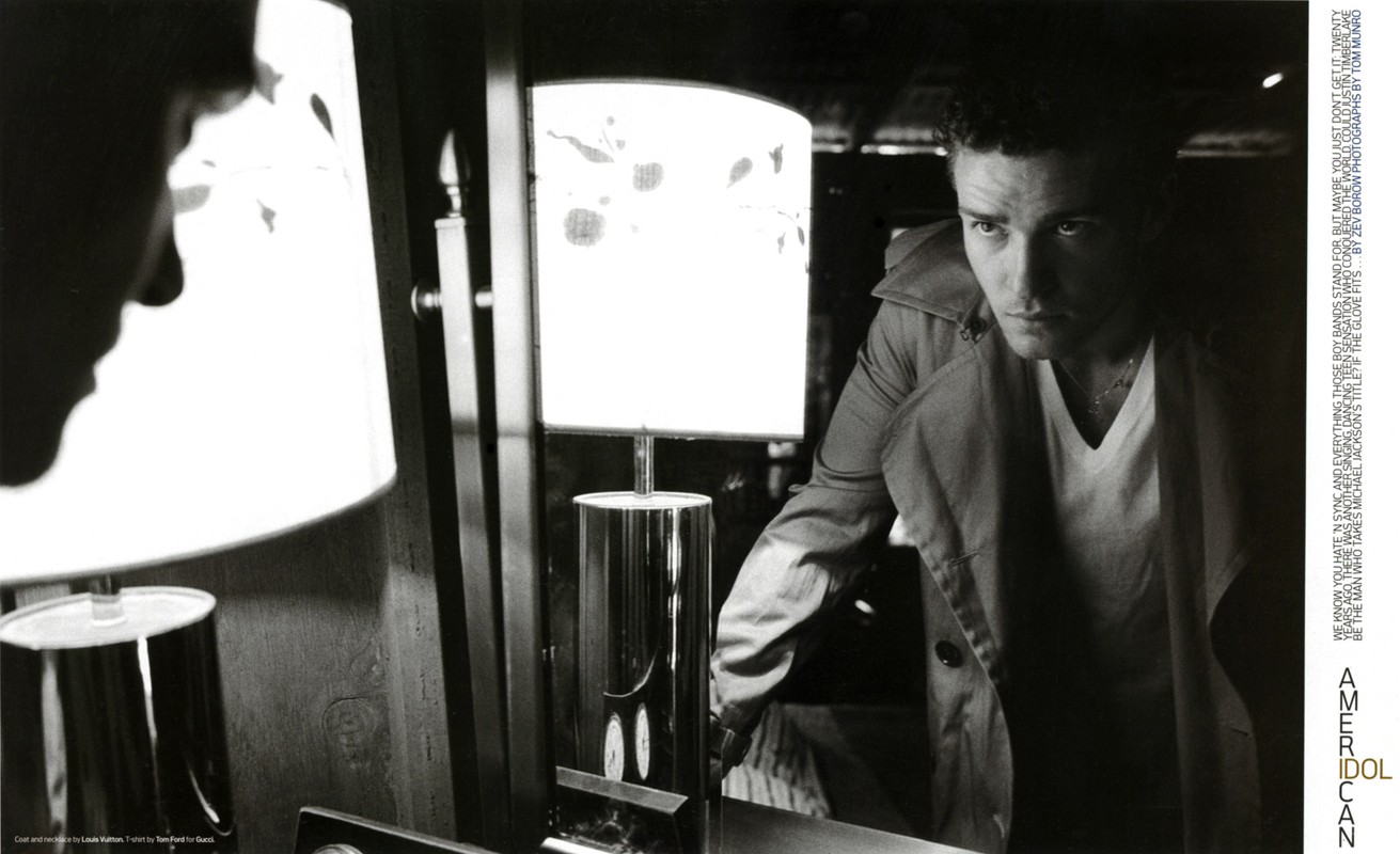 Justin Timberlake in a trench coat and V-neck t-shirt looking into a mirror with a lamp in the foreground