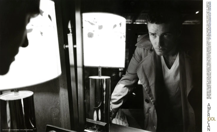 Justin Timberlake in a trench coat and V-neck t-shirt looking into a mirror with a lamp in the foreground