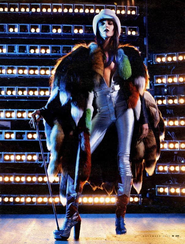A woman in silver jumpsuit and fur coat poses on stage with a cane under blue lighting, fashion-forward and striking.