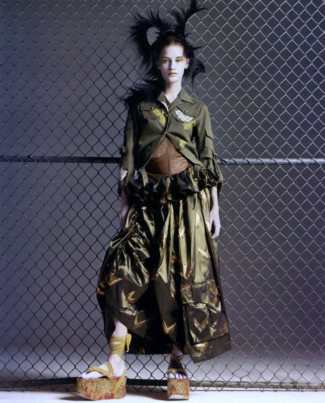 A fashion model strikes a pose in an olive drab military-style jacket and a long skirt, standing on wooden platform shoes with a chain-link fence backdrop.