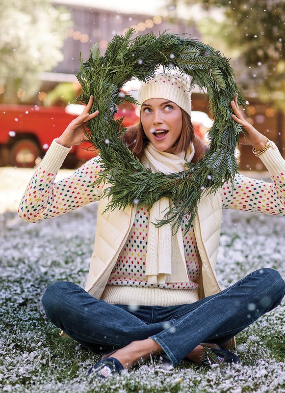 A smiling woman sits cross-legged in a snowy outdoor scene, holding a wreath up to her face, wearing a winter hat, scarf, vest, and jeans.